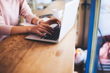 Yüksek açılı tanınmayan bir kadın dizüstü bilgisayarda sıcak içecek ve kağıtlarla birlikte tahta masada oturuyor.