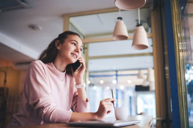 Düşük açılı genç bayan serbest çalışan cep telefonuyla konuşuyor ve masada dizüstü bilgisayar ve bir fincan sıcak içecekle notlar alıyor.