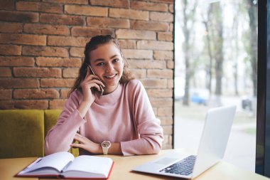 Gündelik giysiler içinde genç pozitif kadın uzaktan çalışan iş saatlerinde telefonla konuşurken pencerenin yanındaki masaya oturmuş not defteri ve modern bilgisayarla kameraya bakıyor.