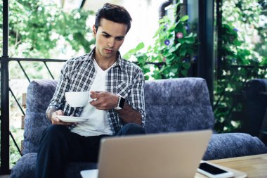 Çay fincanı ile serbest çalışan genç bir erkek sokak kafesinde uzak bir iş yaparken modern dizüstü bilgisayarda web bilgilerine göz atıyor, kahve saatinde webinar video izleme konusunda yetenekli bir bilişim uzmanı.