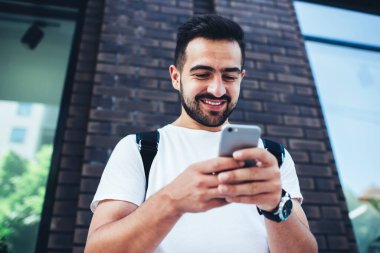 Şebeke takipçileriyle sohbet etmek ve mesajlaşmak için modern mobil cihazda kablosuz internet kullanan neşeli Kafkasyalı turist, şehri ziyaret ederken cep telefonundan internet yayınına göz atan mutlu hipster adam