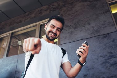Kameraya gülümseyen neşeli erkek turistin yarım boy portresi modern cep telefonuyla sizi işaret ederken dijital akıllı telefon teknolojisi olan Kafkasyalı hippi sokakta mutlu hissediyordu.