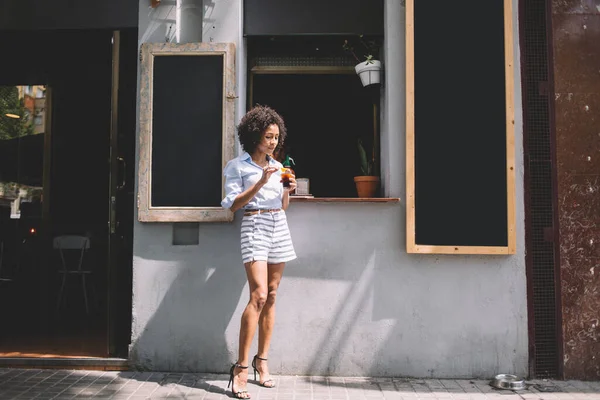 若いアフリカ系アメリカ人の流行の女性がショートパンツやヒールで 日当たりの良い日にカフェの通りの外にカクテルのガラスと巻き毛が立っている — ストック写真