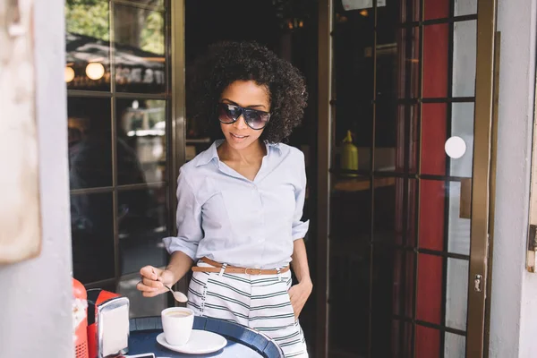 流行のカジュアルな服やサングラスでスタイリッシュな若い民族の女性は 現代のカフェテリアのガラスドアの近くに立ってコーヒーのカップに砂糖を追加します — ストック写真