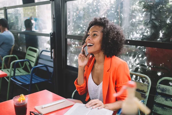 オレンジ色のジャケットで現代アフリカ系アメリカ人女性フリーランスカフェで本を保持しながら 楽観的な笑顔で電話で週末の計画を作る — ストック写真