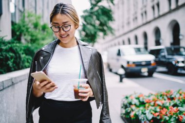 Sosyal medyada mesajlaşmak için mobil uygulama kullanan gülümseyen Asyalı kadın elinde kahve, modaya uygun giyinmiş mutlu Çinli kız açık havada eğlenirken posta kontrol ediyor.