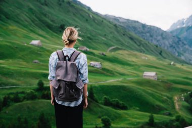 Kırsal kesimdeki yolculukta küçük bir köyün yakınındaki çimenli vadide duran, sarı saçlı, yüzü olmayan kadın turistin arkası.