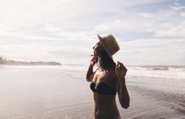 Bikini giyen, şapkalı, gülümseyen ve kumlu sahilde el sallayan mutlu bir kadının yan görüntüsü.