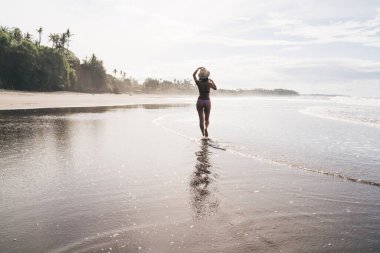 Bikini giyen, şapkalı, deniz kıyısında yürüyen ve yaz tatillerinde güneşli günlerin tadını çıkaran tüm vücudu tanımlanamayan bir kadın.