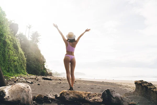 Uzun boylu, tanınmayan, mayo giymiş, şapkalı ince bir kadın tatilde kollarını kaldırmış taşın üzerinde duruyor.