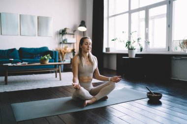 Avrupalı bir kız evde yoga ve meditasyon yaparken gözleri kapalıydı. Sağlıklı yaşam tarzı kavramı. Spor giyim sektöründe genç ve çekici bir bayan. Stüdyo dairenin içi.