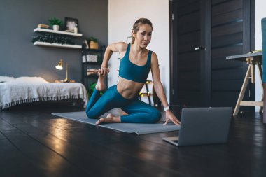 Avrupalı bir kız evdeki spor minderinde dizüstü bilgisayarda bir şeyler izleyip esniyor. Sağlıklı yaşam tarzı kavramı. Spor giyim sektöründe genç ve güzel bir bayan. Stüdyo dairenin içi.