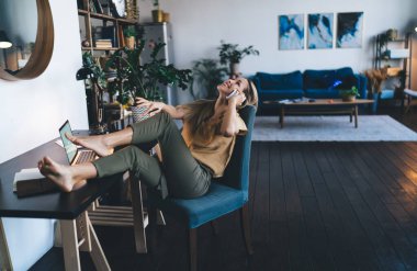 Avrupalı bir kız evde cep telefonuyla konuşuyor. Modern kadın kavramı. Uzaktan veya e-öğrenme fikri. Genç, neşeli bir kadın, masadaki koltukta dizüstü bilgisayarını açmış oturuyor. Stüdyo dairenin içi.