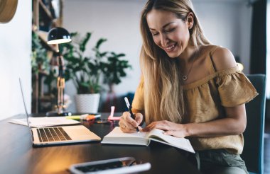 Avrupalı kız not defterine bir şeyler yazıyor. Uzaktan veya e-öğrenme kavramı. Modern kadın fikri işte. Genç gülümseyen bayan, masadaki koltukta açık bir dizüstü bilgisayarla oturuyor. Stüdyo dairenin içi.