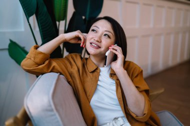 Evdeki koltukta oturup dinlenirken cep telefonuyla konuşan düşünceli Asyalı kadın. Ev hayatı kavramı. Gündelik kıyafetler giyip gözlerini kaçıran çekici bir kız. Geniş daire
