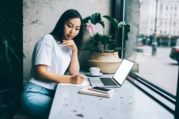 カジュアルな服装の若い民族の女性は 携帯電話やノートパソコンの近くのネット上でリモートで働いてカフェに座っている花と紙の上でコーヒーを飲みながらテーブルで書く — ストック写真