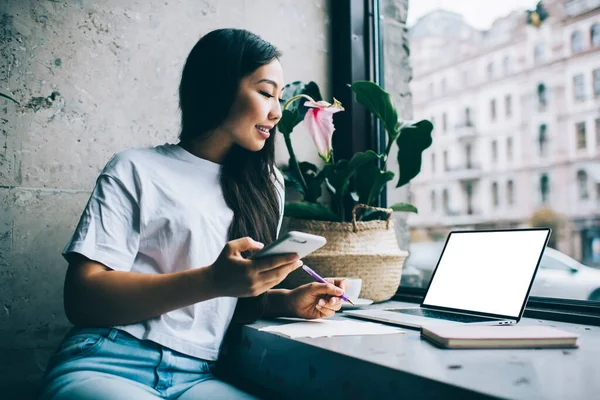 ノートパソコンで宿題をしたり カフェに座ってノートを取ったりしながら スマートフォンでカジュアルな服装で積極的な若いアジアの女性学生 — ストック写真