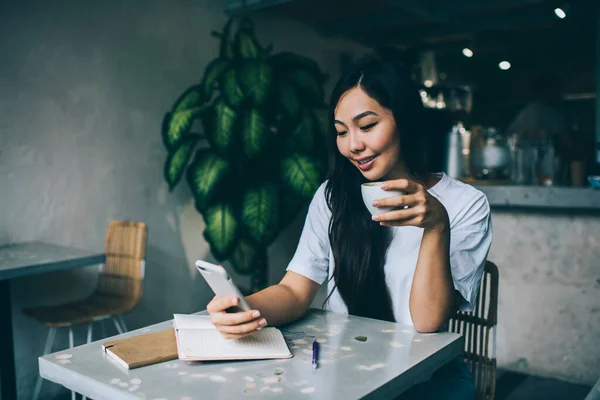 現代的なカフェでノートと一緒に座っている間に携帯電話で熱い飲み物やテキストメッセージを飲む楽しいアジアの女性 — ストック写真