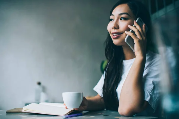 コンテンツのサイドビュー飲み物やメモ帳をテーブルに座っている間に電話を持っているアジアの女性 — ストック写真