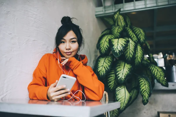音楽を聴きながら スマートフォンでチャットしながら テーブルにもたれてカメラを見て穏やかなアジアの女性 — ストック写真