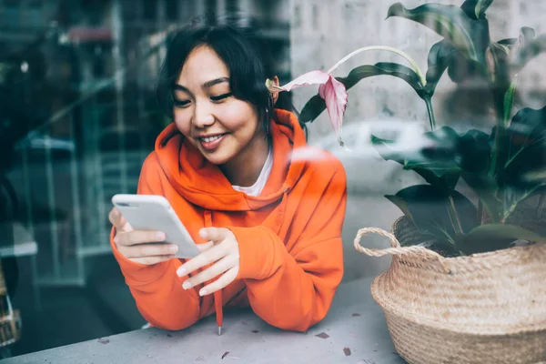 ラウンジゾーンに座り 休憩中に携帯電話でメッセージを送るアジアの女性の笑顔が幸せな窓を通して — ストック写真