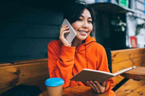 明るいパーカー姿の正の民族女性リモートワーカーは ノートブックでカフェに座っている間にカメラを見て 携帯電話で会話をしながら — ストック写真