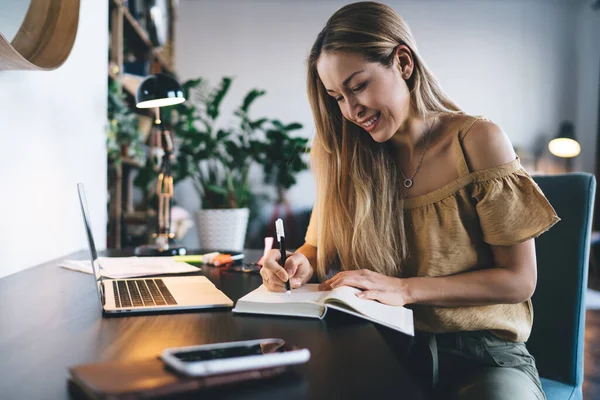 Avrupalı kız not defterine bir şeyler yazıyor. Uzaktan veya e-öğrenme kavramı. Modern kadın fikri işte. Genç, gülümseyen bir bayan açık dizüstü bilgisayarla masada oturuyor. Stüdyo dairenin içi.