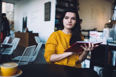 Genç bir kadın, gündelik giysiler içinde elinde kitapla masada oturmuş elinde bir bardak sıcak içecekle kameraya bakıyor.