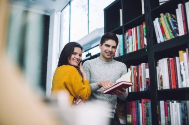 Genç ve neşeli kadın ve erkeğin altında kalın giysiler içinde kitaplıkların yanında açık ders kitabıyla kameraya bakarken...