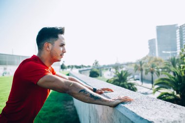 Spor elbiseli, spor elbiseli, koşuya hazırlanan, caddede taş duvara yaslanmış genç bir adamın yan görüntüsü.