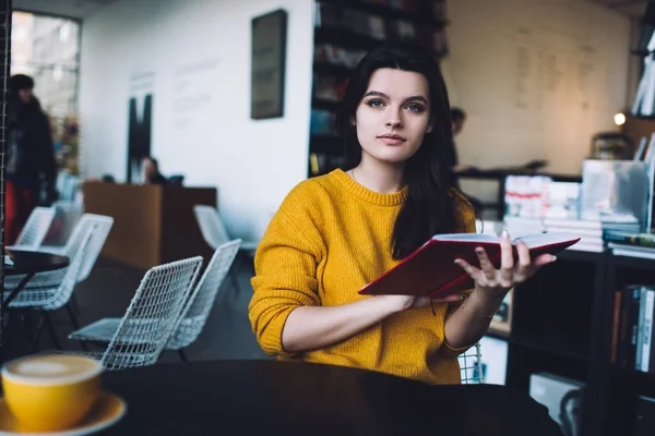 Genç bir kadın, gündelik giysiler içinde elinde kitapla masada oturmuş elinde bir bardak sıcak içecekle kameraya bakıyor.