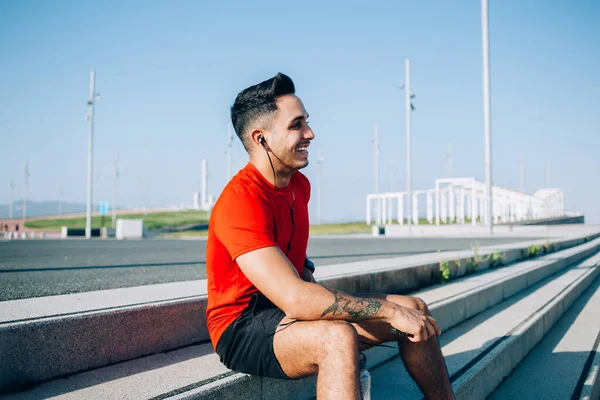 Kırmızı tişörtlü ve mavi şortlu neşeli, kaslı, atletik bir erkek. Güneşli bir günde stadyumda kulaklıkla müzik dinliyor.