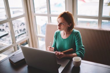 Modern dizüstü bilgisayarla masaya oturup not defterine notlar yazarken gündelik giysiler giymiş ciddi kadın uzaktan çalışan yüksek açılı bir bakış açısı.