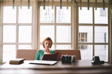 Gündelik kıyafetli genç bir kadın masada oturmuş elinde kitaplarla fotoğraf makinesi ve bir fincan sıcak içecek ve kafede notları kontrol ediyor.