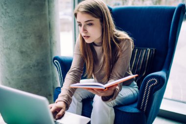 Balıkçı yakalı, kot pantolonlu, elinde kitap tutan rahat koltukta oturmuş internetten bilgi arayan dalgın bir genç kadın.