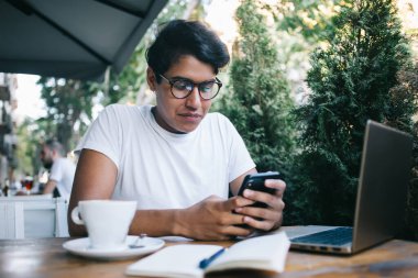 Optik gözlüklü Latin bilişim uzmanı, başvuru için ücretli internet bankacılığı yapıyor. Uzak mesafe için dizüstü bilgisayarla sokak kafesinde oturuyor, cep telefonuyla 4G 'ye bağlanan yetenekli erkek.