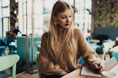Düşünceli, serbest giyinen, kırmızı ruj sürmüş, ahşap masada oturan ve kafedeki deftere not alan bir kadın.