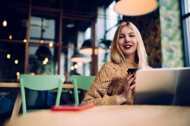 Gülümseyen, modaya uygun bir süveter giyen, dizüstü bilgisayarlı ve akıllı telefonlu, kulaklık takan ve kafeteryanın rahat iç kısımlarına karşı kahve içen genç bir kadın.