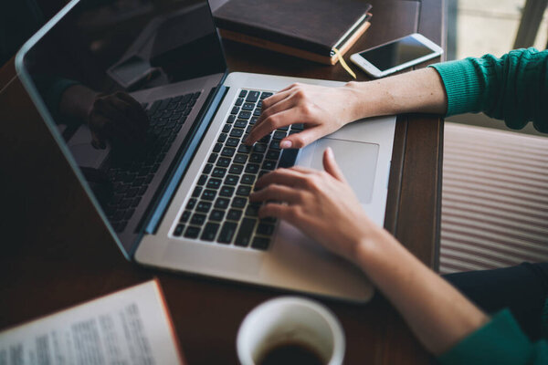 From above of crop unrecognizable female freelancer sitting at wooden table and surfing laptop while working on project remotely in light workplace