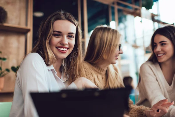 穿着休闲装的积极女性在与朋友一起工作的时候 与剪贴板坐在现代咖啡店里 面带微笑 看着相机 — 图库照片