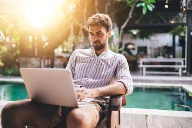 Çizgili tişörtlü mutlu erkek internette geziniyor ve havuz kenarındaki terasta dinlenirken internet üzerinden daktilo yazıyor.