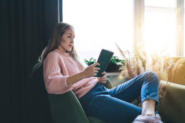 Avrupalı bir kız koltuğunda oturuyor ve evde kitap okuyor. Ev hayatı kavramı. Ev istirahatı ve boş zaman fikri. Gündelik kıyafetler giyen genç ve güzel bir kadın. Stüdyo dairenin içi.