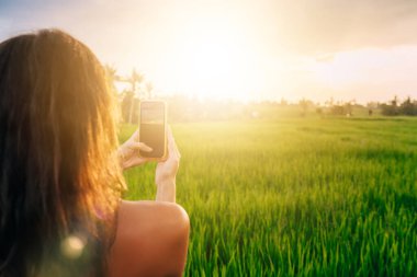 Kadın blogcunun Ubud 'daki Endonezya tatillerinde pirinç tarlalarını fotoğraflamak için cep telefonu teknolojisini kullanışının arka planı, hareketlilik için gezinen internete bağlanan video blogu çeken kadın