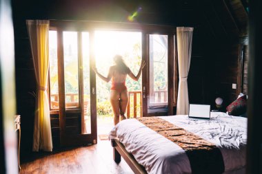 Kırmızı elbiseli zayıf bir kadının, güneşin doğuşunda yatak odasının kapısında dikilirken doğaya hayran olduğu ve temiz hava aldığı arka plan.