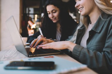 Ciddi kadınlar şık kıyafetler içinde ahşap tezgahta oturuyor ve rahat çalışma alanında dizüstü bilgisayar kullanırken birlikte uzaktan ödev yapıyorlar.