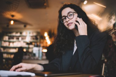 Resmi kıyafetler ve gözlükler içinde karizmatik bir bayan girişimci ahşap masada oturuyor ve kameraya bakarken cep telefonuyla konuşuyor.
