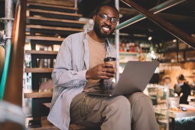 Kahverengi tenli komik bilişim uzmanının portresi, paket kafein içeceği ve bilgisayarı kameraya gülümsüyor, gözlüklü siyah bir blogcu, mesafesinde kahve molası veriyor.