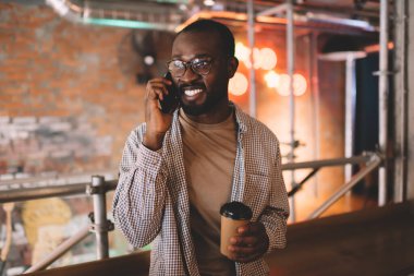Görsel koruma için klasik gözlüklü komik bir erkek kahve molası sırasında pozitif mobil iletişimin keyfini çıkarıyor, mutlu koyu tenli hippi adam telefon görüşmesi için cep telefonu teknolojisini kullanıyor.