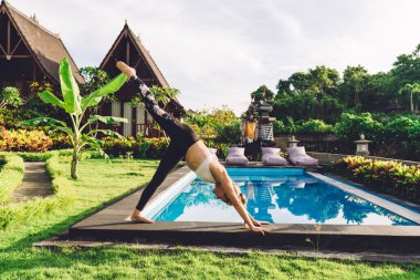 Spor kıyafetli sağlıklı bir kadının havuz başında çalışırken 3 bacaklı köpek pozisyonunda yoga yapması.