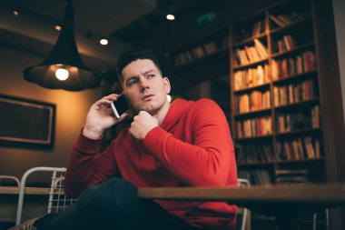 Düşük açılı, kırmızı kapüşonlu genç bir adam. Elleri çene hizasında oturuyor, telefon ediyor ve başka yere bakıyor.